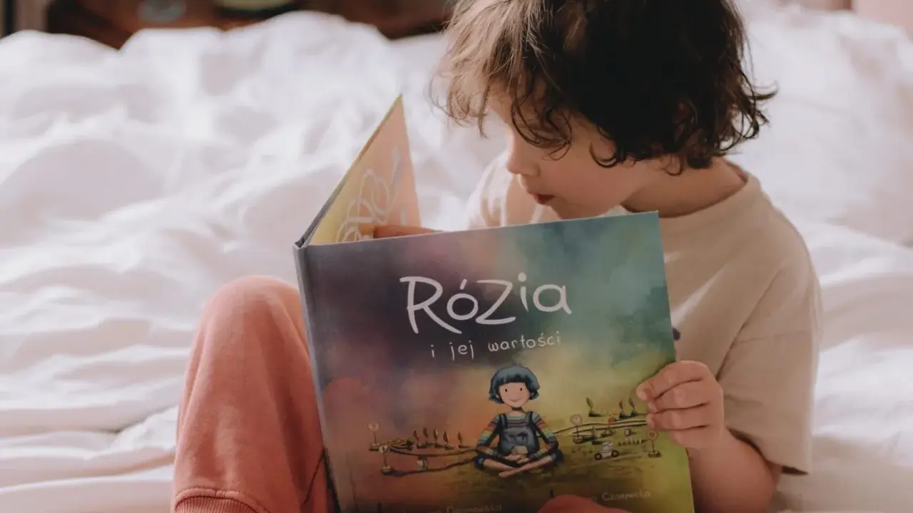 Dziecko czyta książkę "Rózia i jej wartości". Obok leżą karty pracy do nauki dodawania i odejmowania pełnych dziesiątek.