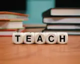 Kolejność wprowadzania liter w przedszkolu mac. Kostki z napisem TEACH na drewnianym blacie, w tle stosy książek.