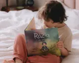 Dziecko czyta książkę "Rózia i jej wartości". Obok leżą karty pracy do nauki dodawania i odejmowania pełnych dziesiątek.