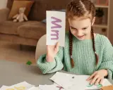Dziewczynka uczy się liter, trzymając kartkę z literą "M". Na stole leżą inne karty z cyfry po śladzie do druku.
