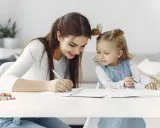 Mama uczy córkę odejmowania do 10. Dziewczynki piszą w zeszytach, a obok leżą kolorowe flamastry.