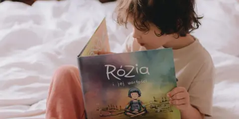 Dziecko czyta książkę "Rózia i jej wartości". Obok leżą karty pracy do nauki dodawania i odejmowania pełnych dziesiątek.