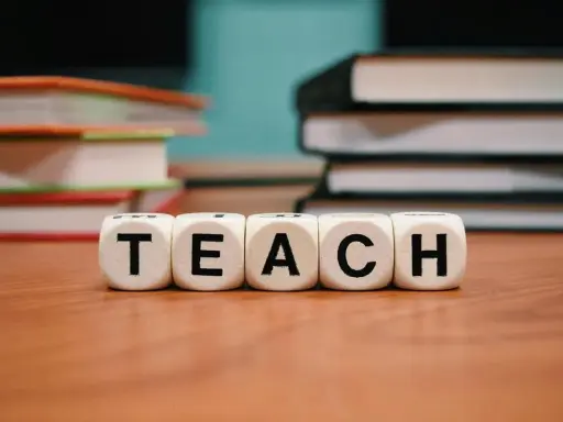 Kolejność wprowadzania liter w przedszkolu mac. Kostki z napisem TEACH na drewnianym blacie, w tle stosy książek.