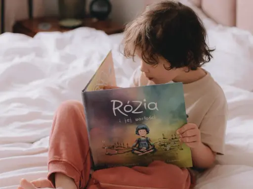 Dziecko czyta książkę "Rózia i jej wartości". Obok leżą karty pracy do nauki dodawania i odejmowania pełnych dziesiątek.